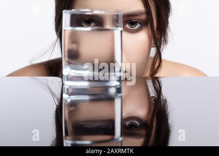 Girl hides her face behind a glass with water. Beauty portrait of young woman at the mirror table. Female on gray background. Stock Photo