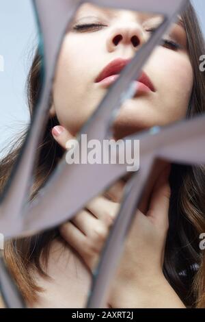 Young woman looks in a broken mirror. Portrait of beautiful female in the mirror shards. Hand on the throat. Stock Photo