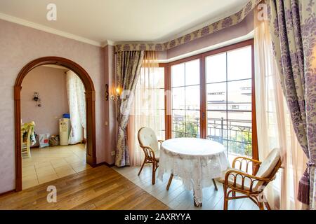 Room with expensive repairs in a classic style. A round wooden table with a white tablecloth and two wicker chairs stand by the large window. Curtains Stock Photo