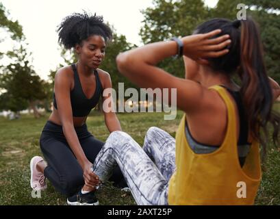 African american friend helping to support her friend doing sit-ups in the park - female friends doing exercise outdoors in the park Stock Photo