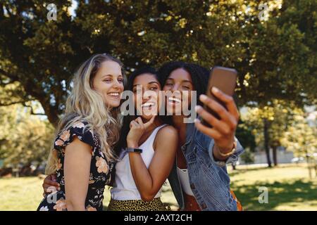 Close-up portrait of happy multiracial smiling group of young woman taking selfie on mobile phone in the park - group of friends laughing and having Stock Photo