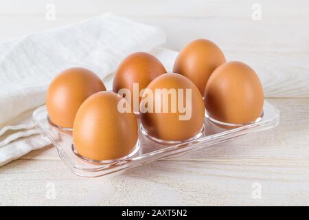 Six raw chicken eggs in a transparent plastic tray on a white wooden table. Half a dozen brown eggs prepared for cooking. Cooking for easter. Stock Photo