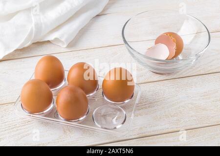 Five raw chicken eggs in a transparent plastic tray and empty eggshell in a glass bowl on a white wooden table. Cooking for easter. Brown eggs. Stock Photo