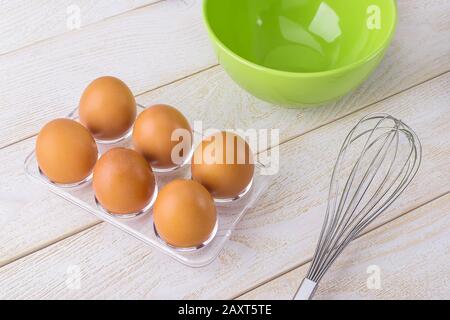 Six raw chicken eggs in a transparent plastic tray, green bowl and stainless steel whisk on a white wooden table. Cooking for easter. Stock Photo