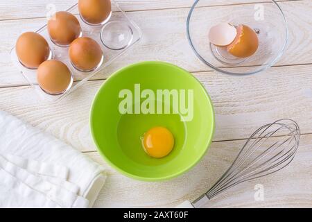 One raw egg in a green bowl, five brown chicken eggs in a transparent plastic tray and whisk on a white wooden table. Cooking for easter. Stock Photo