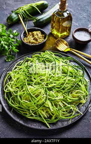 Zucchini pasta, zoodles on a black plate on a grey concrete table with basil sauce pesto, golden fork, spoon and ingredients, vertical view, close-up Stock Photo