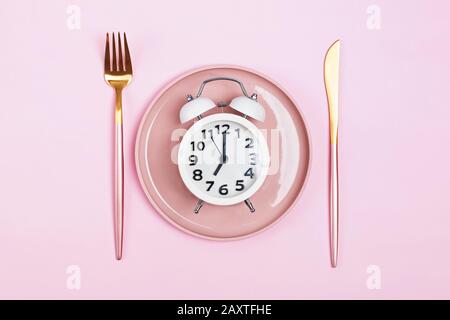 Retro alarm clock on plate with fork and knife isolated on pink background. Stock Photo