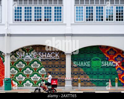 SINGAPORE – 5 JAN 2020 – A young Asian Chinese Zeek delivery driver drives an electric scooter past a Shake Shack restaurant in downtown Singapore Stock Photo