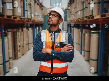 Confident warehouse supervisor in aisle standing with folded hands looking high at shelves with material while wearing orange vest and white hardhat Stock Photo