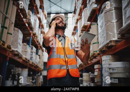 Low angle view of warehouse manager talking over phone while smiling and holding digital tablet standing in aisle with goods Stock Photo