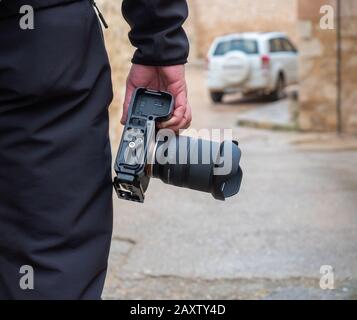 Detail of a man in dark clothes holding a camera in the street with a car in the background Stock Photo