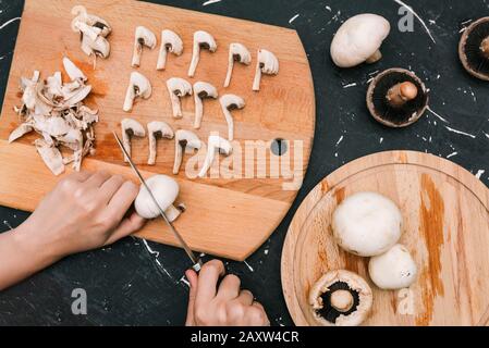 Sliced white champignons on a wooden board. The girl cuts a knife with a knife on a dark background. The process of cooking. Stock Photo