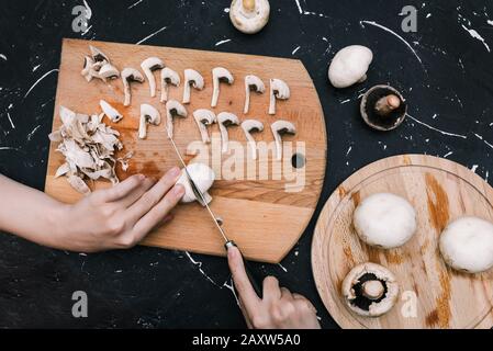 Woman cuts champignons on a cutting board with mushrooms. Hands of a girl with a knife and mushrooms on a dark background. Top view, copy space. Flat Stock Photo