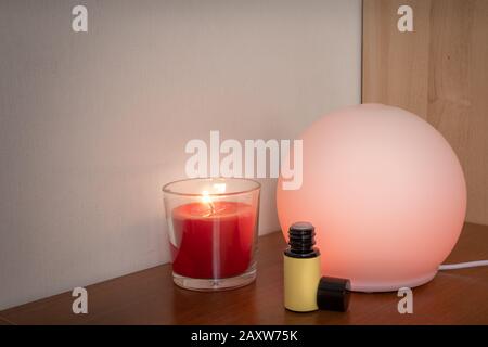 Close up of a candle with flame, fragrance diffuser illuminating the scene with orange light and a bottle of essential oil Stock Photo