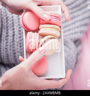 Female hands holding rectangular white plate with delicious pink and beige macaroons on pink and gray background. Top view Stock Photo