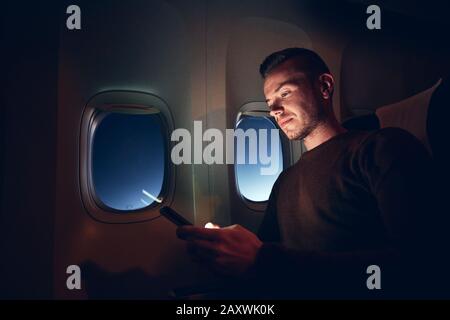 Young man using mobile phone in airplane. Internet connection during flight. Stock Photo