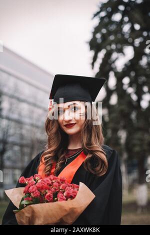 Portrait of young female graduate student in mortarboard holding bouquet of flowers while standing in campus at graduation ceremony Stock Photo