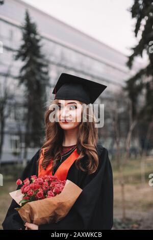 Portrait of young woman in graduation gown holding bouquet of flowers while standing in campus at graduation ceremony Stock Photo