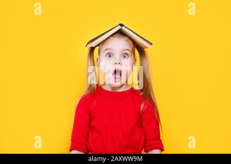 Shocked little girl holding book on her head and looking at camera Stock Photo