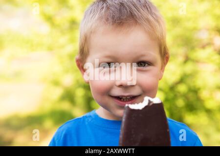 Charming blond smiling boy 5 years old in summer outdoors on hot sunny day eating popsicle in chocolate on stick. Closeup portrait on blurry backgroun Stock Photo