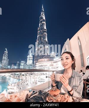 26 November 2019, UAE, Dubai: Happy Asian girl drinking coffee in a cafe with a terrace overlooking the famous skyscraper Burj Khalifa. Nightlife conc Stock Photo