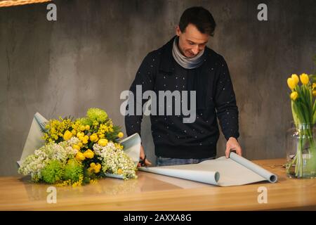 Male make spring bouquet. Man florist wrapping beautiful bouquet of spring flowers in pack paper on the wooden table. Beautiful flower composition of Stock Photo