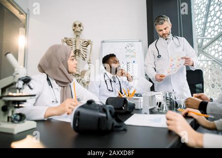Group of multiracial medical students sitting together and studying, listeting to their experienced male professor during the lesson in the classroom Stock Photo