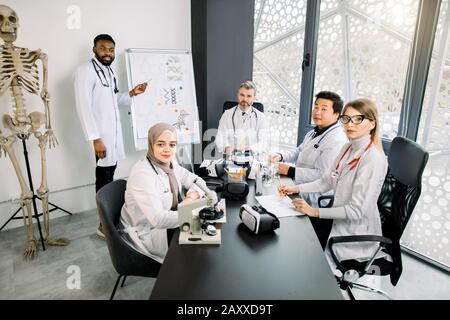 Portrait of middle aged lecturer professor with group of multiethnic students, sitting at the table in modern lab or classroom. African doctor chemist Stock Photo