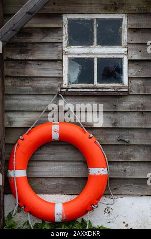 an orange coloured plastic lifebuoy on the side of an old wooden beach hut or cabin with a small square window. Stock Photo