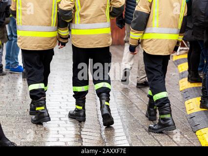 Three anonymous fireman in yellow reflective uniforms and black boots and trousers walking away from the camera on the crowdy street, back side, legs Stock Photo