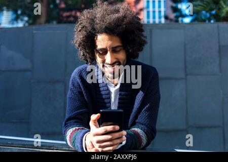 Portrait Young handsome African man using his smartphone with smile while leaning on a fence outdoors in sunny day Stock Photo