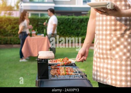 Asian man cooking barbeque grill and sausage for a group of friends to eat party in garden at home. group of friends having outdoor garden barbecue la Stock Photo