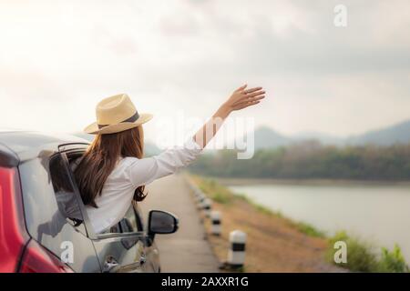 Woman at spree time relaxing and enjoying road trip. Young Asian happines female wearing hat inside compact black car with raise her hand out to the c Stock Photo