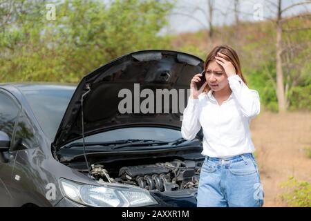 Asian young unhappy woman talking on a cell phone  in front of the open hood  broken down car On Country Road Phoning For Help. Broken Car On The Road Stock Photo