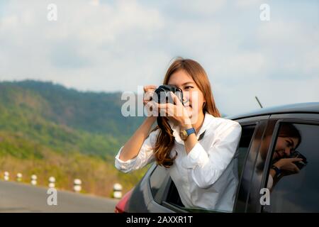 Young Asian Woman tourist taking photo in car with camera driving on road trip travel vacation. Girl passenger taking picture out of window with beaut Stock Photo