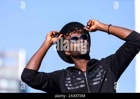 Front view of young black man with sunglasses and hat standing in the street against blue sky while touching hat and looking away in sunny day Stock Photo