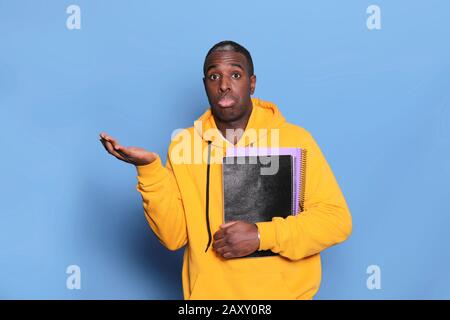 Confused black young man has hesitant expression, holds textbook, isolated over blue background Stock Photo