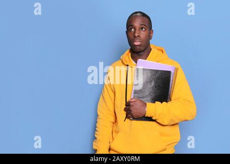 Confused black young man has hesitant expression, holds textbook, isolated over blue background Stock Photo