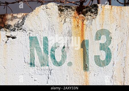 Close-up of a green colored house number 13 painted on a white wall. The paint is peeling off, and rusty barbed wire is leaving stains on the wall. Stock Photo