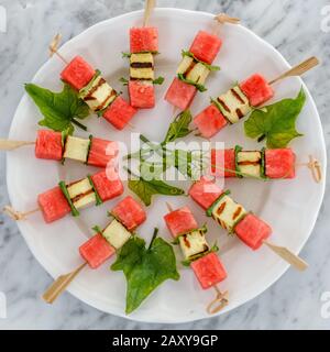 Appetizer with red watermelon, fried hallumi cheese and mint on skewers served on a white ceramic plate. Stock Photo