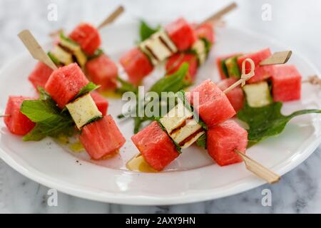 Appetizer with red watermelon, fried hallumi cheese and mint on skewers served on a white ceramic plate. Stock Photo