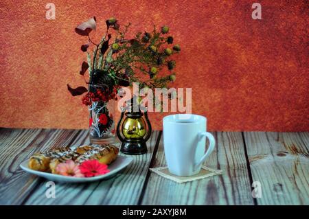 Autumn still life, a tea mug, an old-fashioned lamp, a blackberry and a plate with three eclairs stand on a table against a red wall. Close-up. Stock Photo