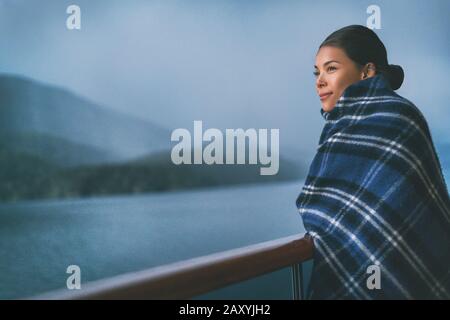 Cruise ship passenger on Alaska travel vacation enjoying scenery at dusk on suite balcony deck with wool throw in cold weather. Asian tourist woman relaxing on summer holiday cruising adventure. Stock Photo