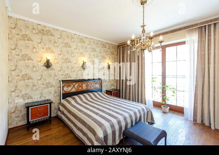Classic Bedroom with beige Wallpaper. A large bed with a bedspread with black stripes. White Bedside tables with red drawers. the lamp on the walls. Stock Photo