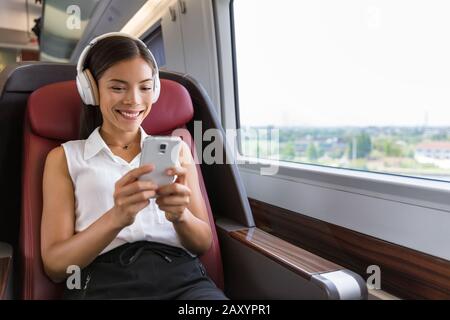 Modern people city lifestyle. Young urban woman using phone app and wireless headphones to listen to music or play video games online. Asian girl enjoying train travel in business class seat. Stock Photo