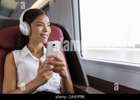 Woman on train listening to music on smartphone. People lifestyle. Young urban businesswoman using phone app, wireless headphones to listen to audiobook. Asian girl enjoying travel in business class. Stock Photo