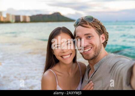 Selfie couple on Waikiki beach taking pictures with smartphone during night out walk on beach summer vacations in Honolulu, Hawaii. Travel destination. Young people having fun on hawaiian holidays. Stock Photo