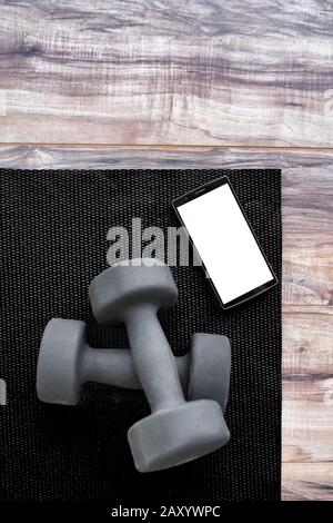 Gym dumbbells on fitness mat showing phone app. Weights on exercise yoga mat and smartphone for health progress tracking mobile app. Phone showing copy on screen for exercising videos online. Stock Photo