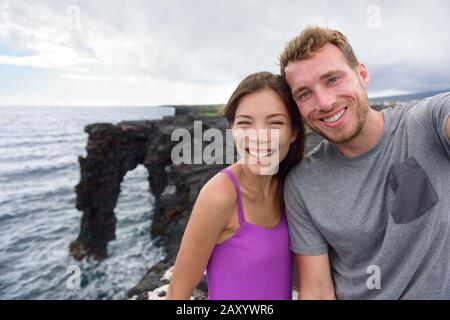 Selfie couple on nature travel Hawaii vacation. Young people taking phone picture at the Holei Sea Arch, tourist attraction on Big Island at the Volcanoes National park. Stock Photo