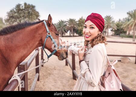 Happy Asian girl in turban touching and petting cute arabian horse. Hobby and friendship between animal and human concept Stock Photo